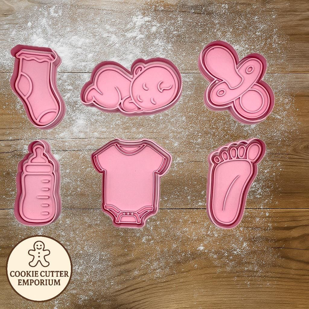 Set of pink baby-themed cookie cutters on a wooden surface with a rolling pin and checkered towel.
