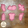 Set of pink baby-themed cookie cutters on a wooden surface with a rolling pin and checkered towel.