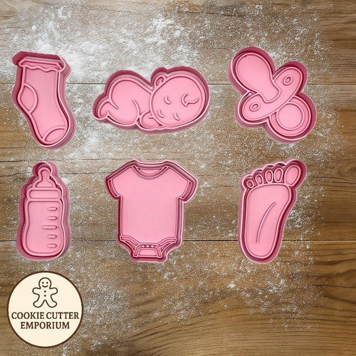 Set of pink baby-themed cookie cutters on a wooden surface with a rolling pin and checkered towel.