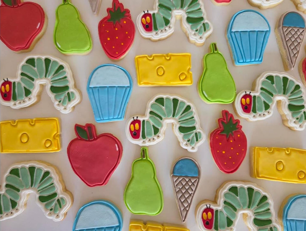 Colorful cookies shaped like a caterpillar, apple, cheese, and other food items on a light background.