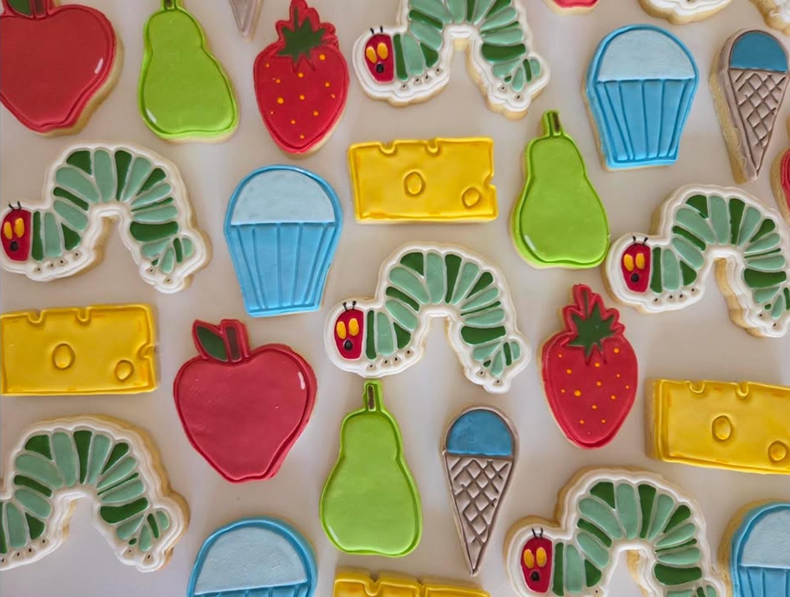 Colorful cookies shaped like a caterpillar, apple, cheese, and other food items on a light background.
