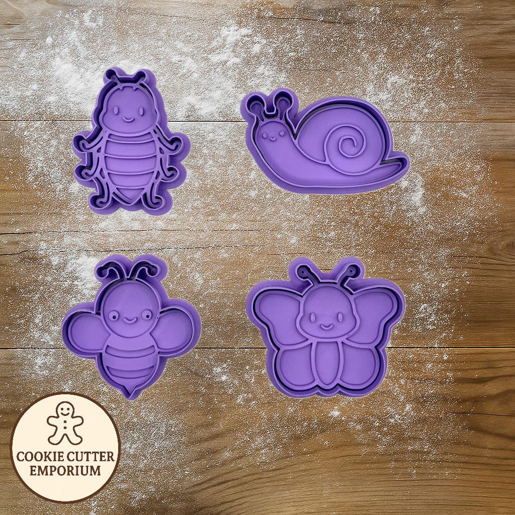 Purple cookie cutters shaped like a caterpillar, snail, bee, and butterfly on a wooden surface with a rolling pin and checkered towel.