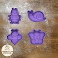 Purple cookie cutters shaped like a caterpillar, snail, bee, and butterfly on a wooden surface with a rolling pin and checkered towel.