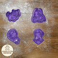 Purple cookie cutters with cartoon designs on a wooden surface with a rolling pin and checkered towel.