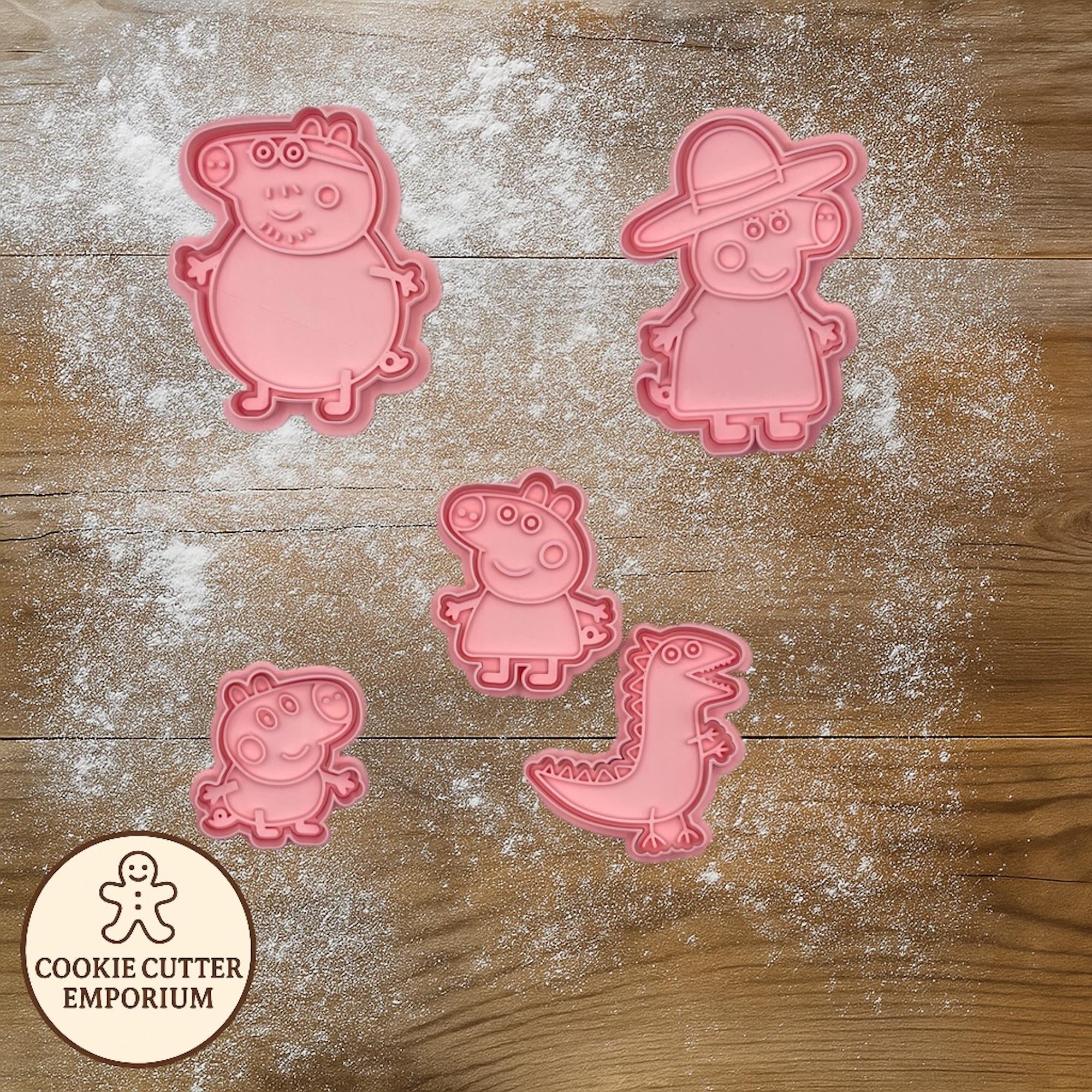 Pink cookie cutters shaped like cartoon characters on a wooden surface with a rolling pin and checkered towel.