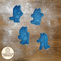 Set of blue cookie cutters shaped like cartoon characters on a wooden surface with a rolling pin and checkered cloth.