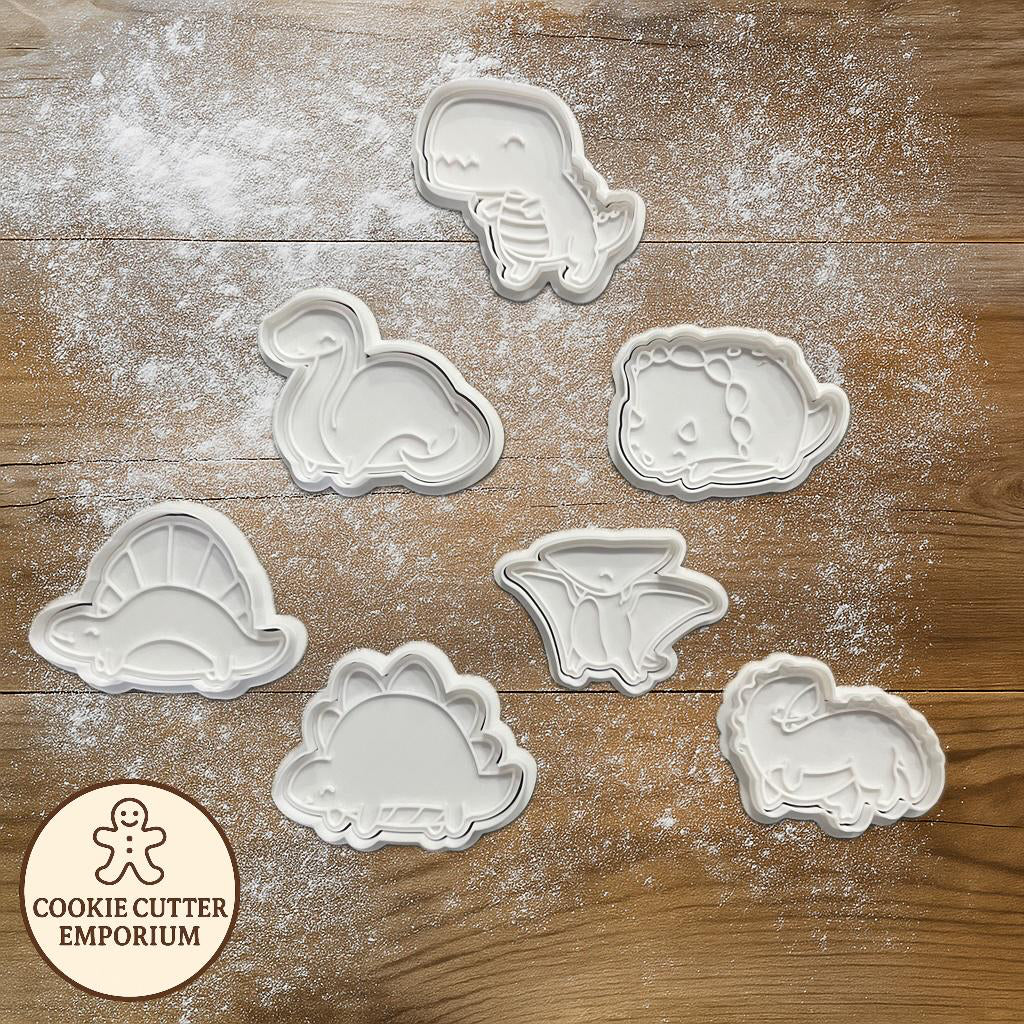 Set of dinosaur-shaped cookie cutters on a wooden surface with a rolling pin and checkered towel.